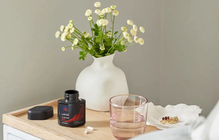 White vase with flowers on a wooden tray with a glass and a bottle of MitoQ Pure.