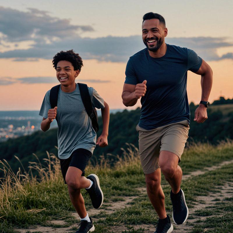 A man and a boy running together on a grassy path at sunset, both wearing athletic t-shirts and shorts. The man wears a dark navy t-shirt and beige shorts, while the boy wears a heather gray t-shirt and black shorts.